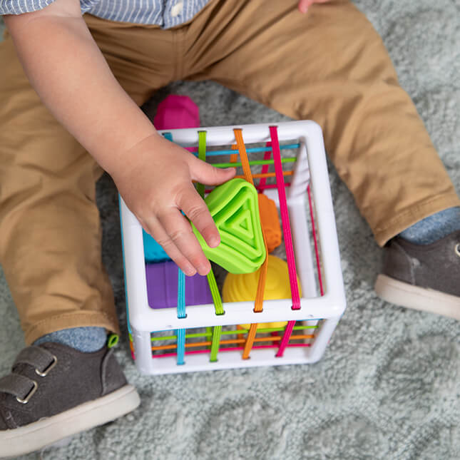 Fat Brain Toys InnyBin – sensory shape‑exploration cube with six chunky, textured blocks (cube, diamond, sphere, triangle, flower, logo) and elastic bands for frustration‑free fine motor play