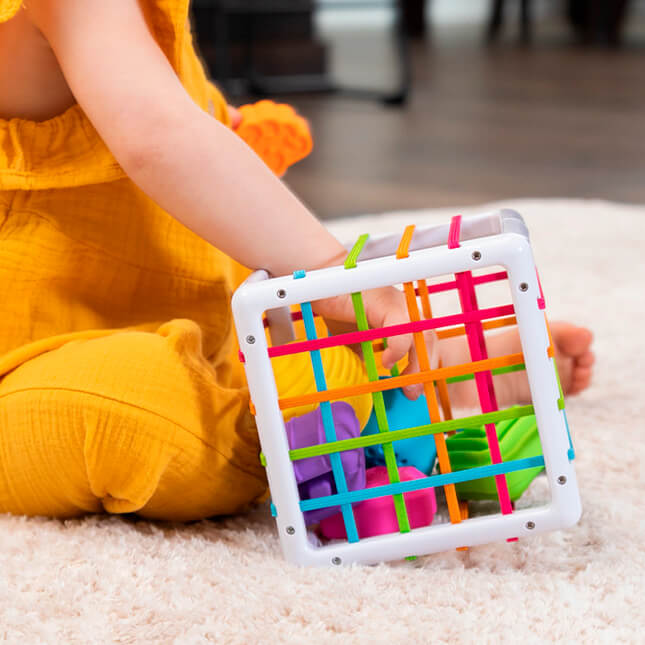 Fat Brain Toys InnyBin – sensory shape‑exploration cube with six chunky, textured blocks (cube, diamond, sphere, triangle, flower, logo) and elastic bands for frustration‑free fine motor play