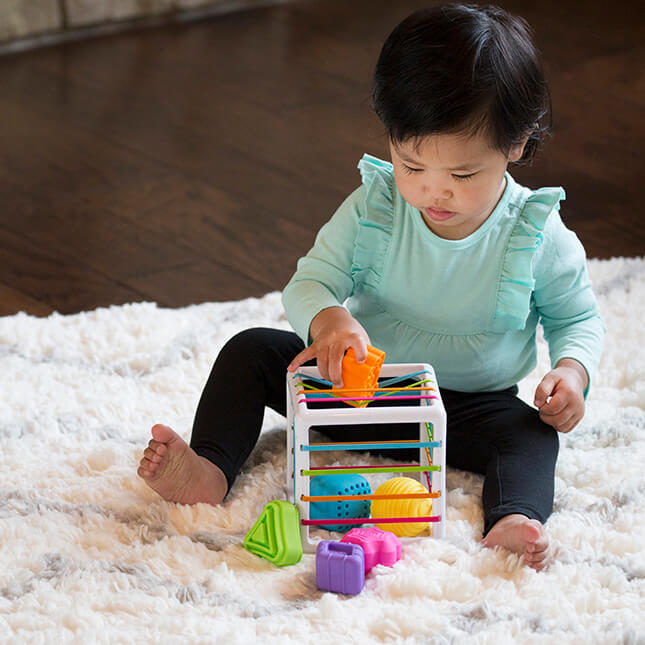 Fat Brain Toys InnyBin – sensory shape‑exploration cube with six chunky, textured blocks (cube, diamond, sphere, triangle, flower, logo) and elastic bands for frustration‑free fine motor play