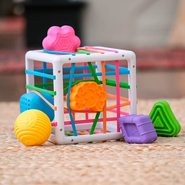 Fat Brain Toys InnyBin – sensory shape‑exploration cube with six chunky, textured blocks (cube, diamond, sphere, triangle, flower, logo) and elastic bands for frustration‑free fine motor play